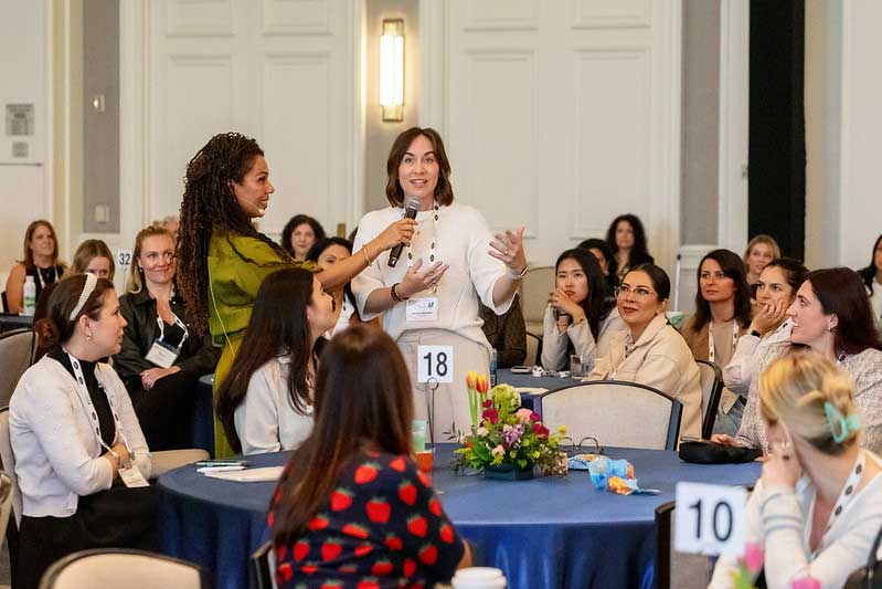 Women participate in a conference discussion, with one speaker holding a microphone beside a standing attendee in a green blouse.